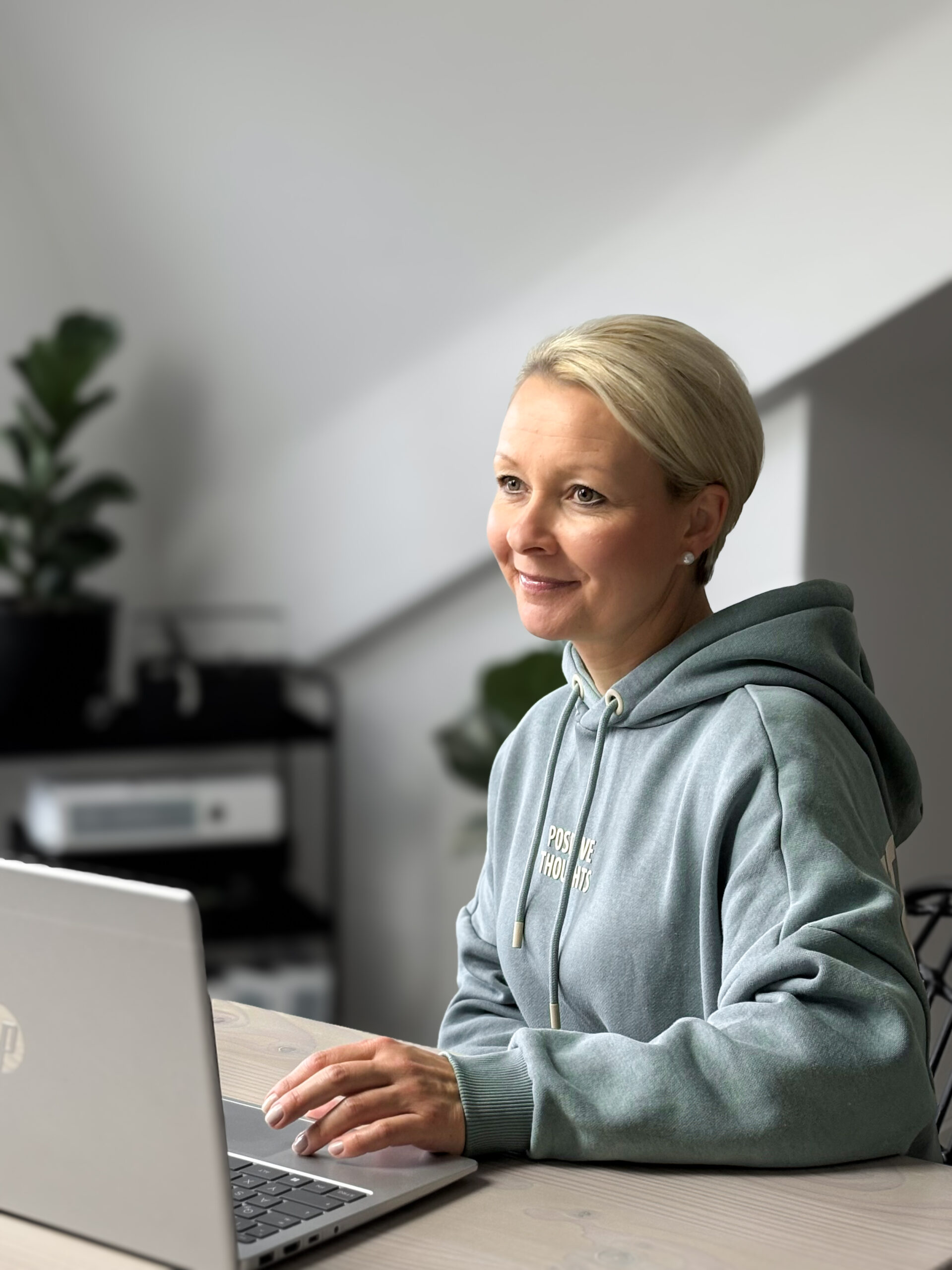 Frau am Laptop in gemütlichem Raum - lächelnd, konzentriert und in einem hellen Hoodie.