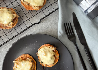 Mehrere überbackene Süßkartoffel-Scheiben mit einer herzhaften Belagsschicht aus Zwiebeln und Mozzarella liegen auf einem schwarzen Teller und einem schwarzen Gitterrost. Daneben sind eine schwarze elektrische Salz- oder Pfeffermühle mit der Aufschrift „KoRo“, schwarzes Besteck und eine hellgraue Stoffserviette auf einem hellgrauen Tisch platziert.