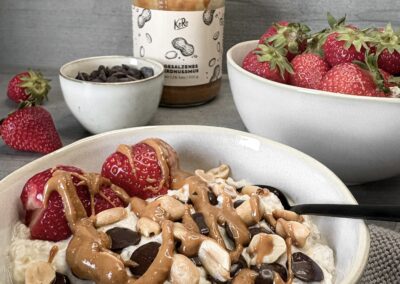 Ein cremiger Porridge in einer weißen Schale, getoppt mit frischen Erdbeeren, knackigen Erdnüssen, dunklen Schokodrops und einem Klecks Erdnussmus. Im Hintergrund stehen weitere Schalen mit Erdbeeren und Schokolade sowie ein Glas Erdnussbutter – alles liebevoll arrangiert auf grauem Untergrund mit einem gestrickten Tuch.